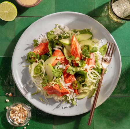 A plate with a prepared meal of salmon and a cilantro slaw salad with apples and cashews on top of a bed of rice.